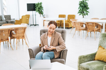 Adult smiling brunette business woman forty years with long hair in stylish beige suit and jeans working on laptop at the public place, open space office