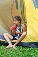 Cute happy caucasian girl is sitting near a camp tent and drinking tea in the morning forest, vertical
