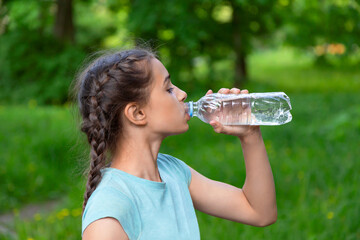 Brunette caucasian girl with braided hair is drinking water from a plastic bottle in green summer park