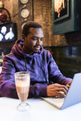 Young African American male freelancer in stylish hoodie sitting at table and typing on netbook and...