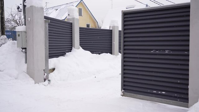 View of an automatic gate closing at snowy winter day. Modern design motorized automatic driveway gate, fence gate of courtyard of residential building in winter.