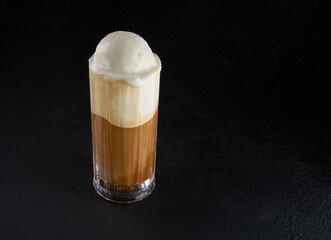 A modern, lactose free coffee drink or cocktail with ice cream and almond milk. Macchiato in a tall glass on a dark background. Cafe beverage layered with milk in a high drinking glass. Copy space