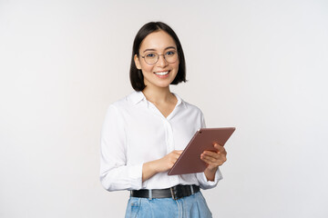 Image of young asian woman, company worker in glasses, smiling and holding digital tablet, standing over white background - Powered by Adobe
