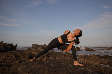 Slim Asian woman practicing yoga on the beach. Standing pose Parivrtta Parsvakonasana. Hands in...