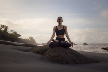 Meditation yoga on the beach. Asian woman sitting on the rock in Lotus pose. Padmasana. Hands in gyan mudra. Yoga retreat. Closed eyes. Healthy concept. Copy space. Mengening beach, Bali