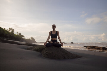 Morning meditation and yoga on the beach. Asian woman sitting on the rock in Lotus pose. Padmasana. Hands in gyan mudra. Yoga retreat. Healthy concept. Copy space. Mengening beach, Bali