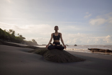 Fototapeta premium Morning meditation and yoga on the beach. Asian woman sitting on the rock in Lotus pose. Padmasana. Hands in gyan mudra. Yoga retreat. Healthy concept. Copy space. Mengening beach, Bali