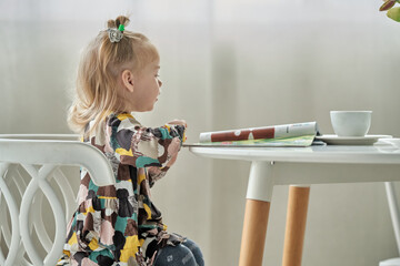 Cute little girl with ponytails on her head reading a magazine sitting on a chair at the table.