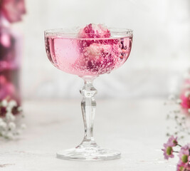 Close up of sparkling pink drink with rose petals in champagne glass at white table with blurred...