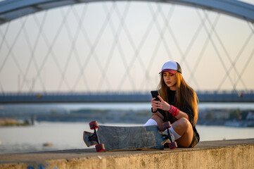 Young woman sitting near the river and using smartphone after longboard ride