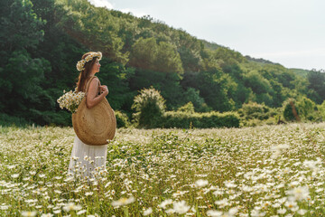 A young woman in a white sundress, a wreath of daisies with a large wicker bag on her shoulder is walking through a field of daisies, against the background of the forest, the sunset light