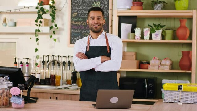 In a cute coffee shop a black charismatic man barista typing something on the laptop then looking to the camera and posing he smiling cute