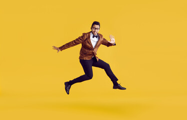 Fototapeta na wymiar Happy man jumping in studio. Cheerful funny young ethnic guy wearing leopard jacket, bowtie and glasses jumping in air, looking at camera and waving hello isolated on vibrant yellow background