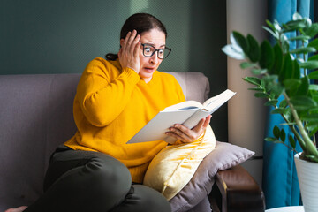 Frightened scared middle-aged woman in eyeglasses reading horror novel, thriller or scary stories...