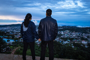 young couple watching downtown city view with dramatic cloudy sky at evening from mountain top