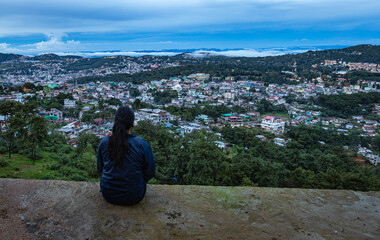 Naklejka premium young girl watching downtown city view with dramatic cloudy sky at evening from mountain top