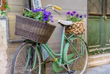 Fototapeta na wymiar Finalborgo, Finale Ligure, Italy. May 5, 2021. In a small street in the center an old green bicycle leaning against a wall with wicker baskets with flower pots inside.