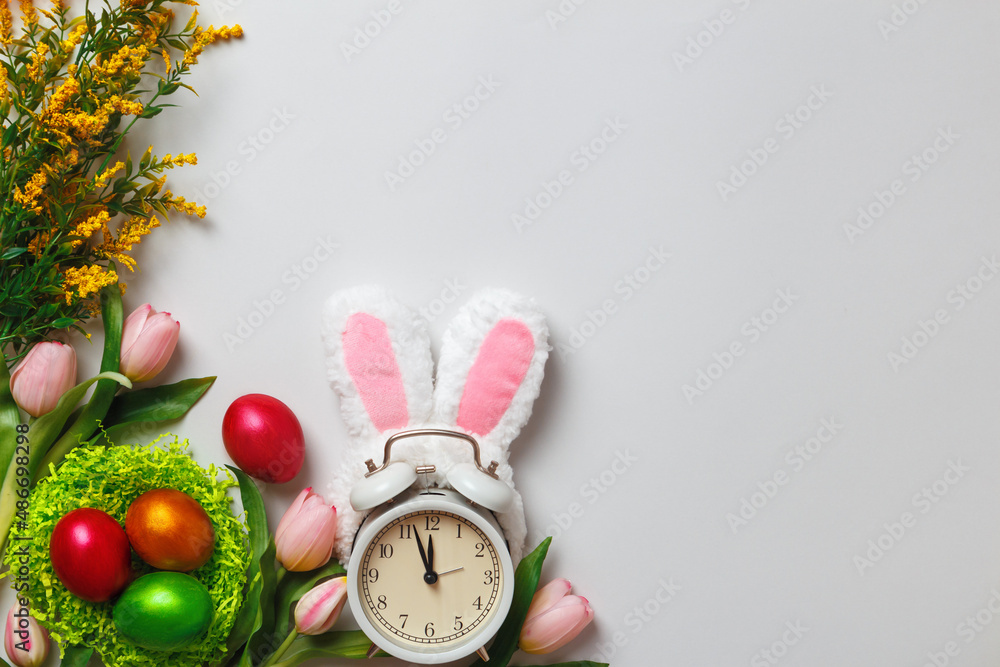 Poster Easter composition. Multicolored pearl eggs in a nest alarm clock rabbit ears flowers tulips on a gray background with copy space.