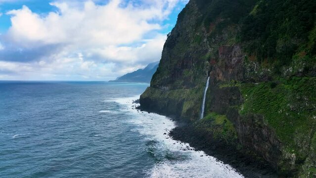 The Atlantic ocean seaside with a waterfall. The seashore waterfall on Madeira. Ocean rocky banks on Madeira island. Green mountains. Beautiful seascape with a waterfall from the rocks. Portugal
