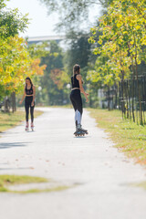 Fototapeta premium Rearview of two young fit women on roller skates riding outdoors on urban street in the park.