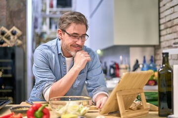 Handsome older man wearing eyeglasses, cooking at home in the kitchen, following recipe in online cookbook app, using tablet.