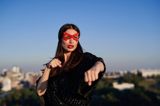 Female power, protest, women rights concept. Brunette superhero girl in black dress and red face mask make boxing using hands on a camera with urban city background. High quality image