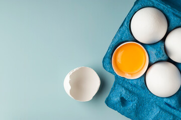 White chicken eggs in eco-packaging on a blue background. Broken egg with yolk in the shell. Farm...