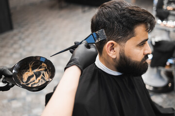Professional barber dying male client hair in barbershop. Hairdresser applies dye to the client's hair with a brush