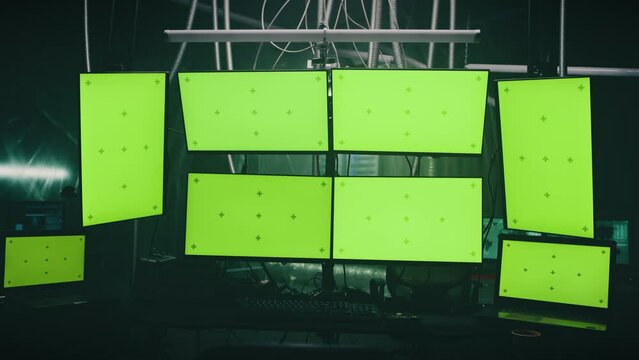 Computers and multiple monitors with chromakey green screen for cyber crimes and cyber attacks on a desk in a hackers dim room with lamps on base. 