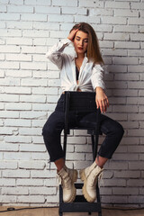 Portrait of a beautiful sensual and sexy blonde girl in a blue dress sitting on a chair against a white brick wall in the Studio