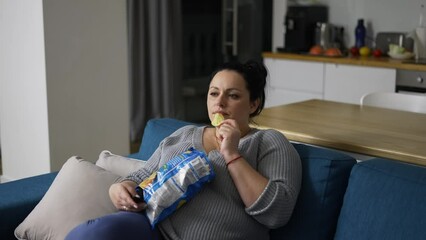 Overweight woman sitting on couch at home, watching tv and eating potato chips