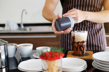Asian barista young man pouring fresh Chocolate art in a plastic glass of Cappuccino iced coffee for according to order at counter bar in the morning at modern cafe.