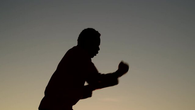 Silhouette of african-american man training to boxing outdoors against background sky, side view. Man trains martial arts. Martial arts concept