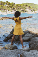 Beautiful female Latina wearing a yellow dress standing on a rock on the seashore