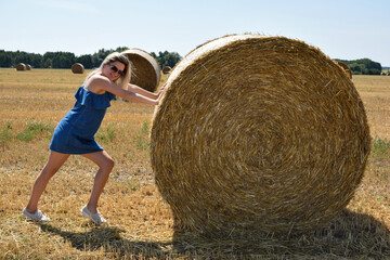 Naklejka na ściany i meble Young woman smiling jokingly pushes a round haystack in a cereal field