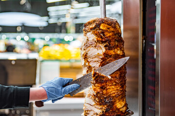 hands holding a knife, cutting slices meat from a vertical skewer. Doner Kebab in market