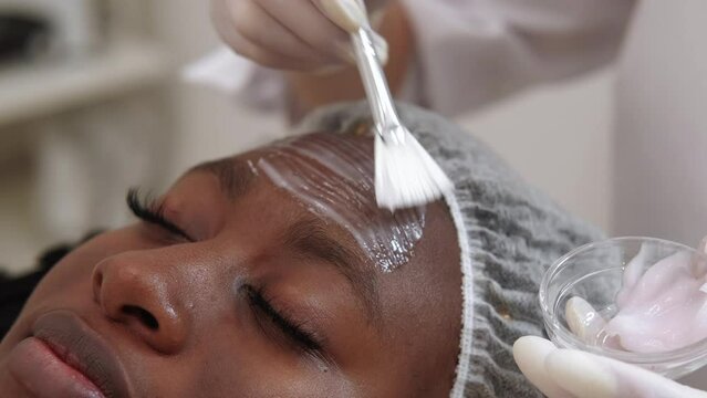 Beauty cosmetology. Skin moisturizing. Facial procedure. Closeup of dermatologist hand applying cosmetic mask organic product with brush on woman client face skin at salon.
