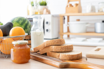 Selective focus on sliced whole wheat bread on wooden tray with apricot jam in jar, bottle of fresh milk, oranges, avocado and watermelon for juices. Breakfast ingredients are on kitchen table at home