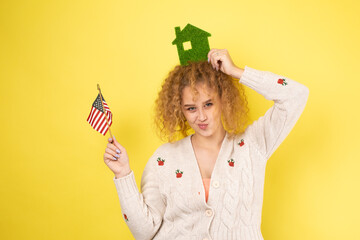 A young girl holds a model of a green house in her hands and an American flag. The concept of buying an eco house.