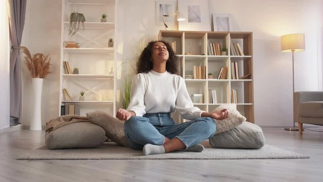 Home meditation. Yoga relax. Wellness leisure. Peaceful calm serene woman meditating in lotus pose on floor with pillows at cozy living room.