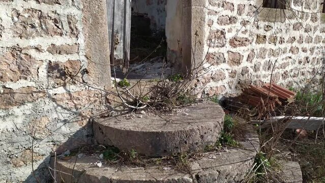 Ruin of old, abandoned and dilapidated stone house - exterior. Concrete steps, crooked wooden door and empty window holes on the front wall of derelict building in village deserted due to rural flight