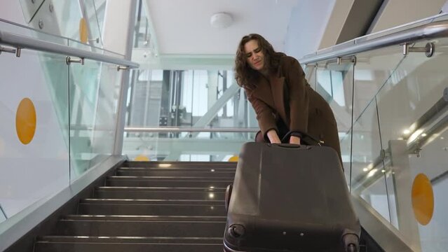 Slow motion footage of young elegant woman carrying heavy suitcase up the stairs at the airport. The concept of traveling with heavy luggage, being late for a flight