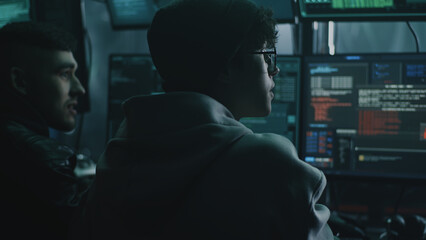 Young male hacker in glasses looking at screen and talking with associate while performing cyber...