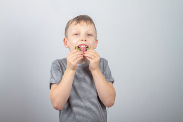 A caucasian boy eats a green kiwi fruit and laughs and smiles on a gray background