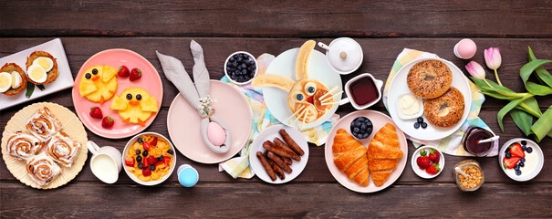Fun Easter breakfast or brunch table scene. Above view on a dark wood banner background. Bunny pancake, egg nests, chick fruit and a selection of spring food items.