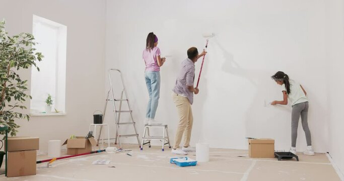 A young father and daughters renovate an apartment in the absence of his wife, surprise, they hold rollers with white paint and precisely painting the walls, a girl climbs a ladder