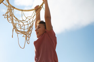 Teenage boy having fun and hanging on basket hoop in yard.