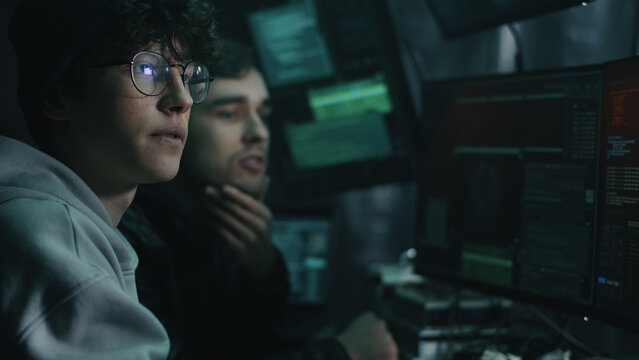 Pan left rack focus of young men discussing hacker attack while sitting at table and gaining access to database in dim room