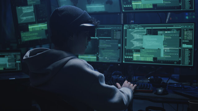 Young cybercriminal in augmented reality glasses hacking secret data in hideout equipped with computers with software and talking about cyber attack