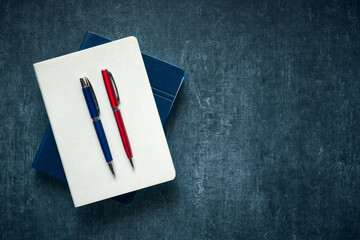 Two pens, blue and red, resting on a white closed notebook, dark grunge background, mockup